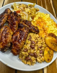 Delicious jerk chicken wings with cabbage, macaroni, rice, and plantains on a plate.