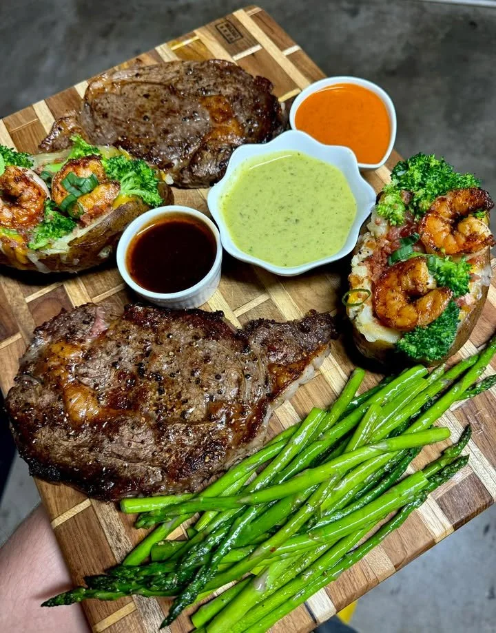 Ribeyes served with loaded shrimp, bacon-baked potatoes, and asparagus on a wooden board.